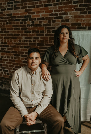 Man sitting on a stool with a woman standing behind him against a brick wall.